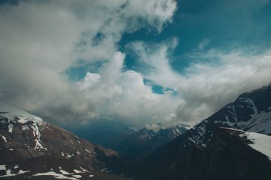 Bulutlu bir gökyüzünün altında karla kaplı kayalık bir dağ manzarası. Tepeleri kar ve bulutlarla kaplı dağlık bir arazi. Dağların tepelerini kaplayan sihirli bulutlar