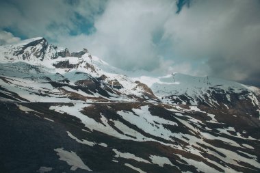 Bulutlu bir gökyüzünün altında karla kaplı kayalık bir dağ manzarası. Tepeleri kar ve bulutlarla kaplı dağlık bir arazi. Dağların tepelerini kaplayan sihirli bulutlar