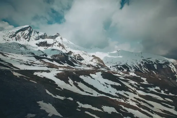 Bulutlu bir gökyüzünün altında karla kaplı kayalık bir dağ manzarası. Tepeleri kar ve bulutlarla kaplı dağlık bir arazi. Dağların tepelerini kaplayan sihirli bulutlar