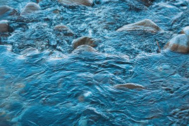 Nehirde su yüzeyi. Kristal berrak su parlak taşların üzerinde dans eder, sakin bir ormanda hafif dalgalanmalar yaratır. Güneş ışığı yaprakların arasından süzülerek doğanın güzelliğinin dingin atmosferini arttırır.