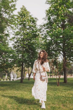 Canlı bir parkın ortasında, bir kadın dalgalı bir çiçekli elbiseyle güvenle yürür, saçları dalgalar halinde dalgalanır. Güneş ışığı ağaçların arasından süzülüp, bu huzurlu ortamda neşeli varlığını vurgular.