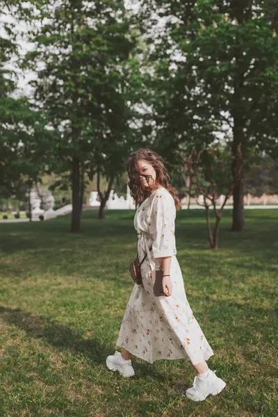 Yeşillik ve ağaçların arasından süzülen güneş ışığıyla çevrili canlı bir parkta zarif bir şekilde gezinen bir kadın.