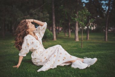 Sakin bir parkta, genç bir kadın yumuşak çimenlerde dinleniyor, beyaz çiçekli elbisesi yavaşça akıyor. Güneş ışığı dalların arasından süzülüp, barışçıl bir ortam yaratır, yansıma için mükemmeldir.