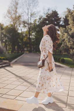 Neşeli bir kadın canlı yeşillikler arasında zarif bir şekilde yürür, sıcak güneş ışığında güneşlenir. Çiçekli elbisesi ışığı yakalıyor, bahçenin huzurlu atmosferini güçlendiriyor.