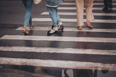 Islak bir yaya geçidinde kalabalık bir caddede insanlar yürüyor. Su onların farklı ayakkabılarını yansıtır ve yağmurlu bir öğleden sonra şehirde canlı bir sahne yaratır.