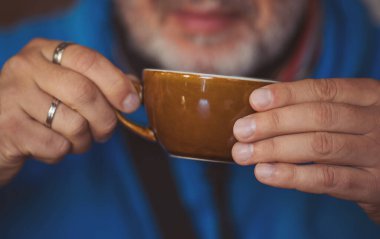 Bir adam, kahve bardağını iki eliyle tutarak rahat bir kafe ortamında içkisinin tadını çıkarıyor. Yumuşak ışıklandırma anın samimi duygusunu arttırır.