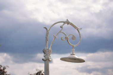 Süslü bir sokak lambası gururla bir parkta bulutlu bir gökyüzünün altında durur. Karmaşık detaylar ve zarif kıvrımlar göstererek çevredeki yolu aydınlatır.