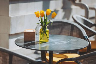Güneşli kafe masası, parlak sarı lalelerle süslenmiş vazoda öğleden sonraları için ortam yaratıyor.