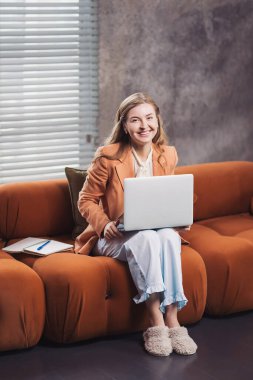 Gülümseyen girişimci evde dizüstü bilgisayarla çalışırken aynı zamanda rahat rahat bir şekilde rahat bir yerde rahat rahat oturuyor. Evden çalışan, iş ceketli, pijama pantolonlu ve terlikli bir kadın.