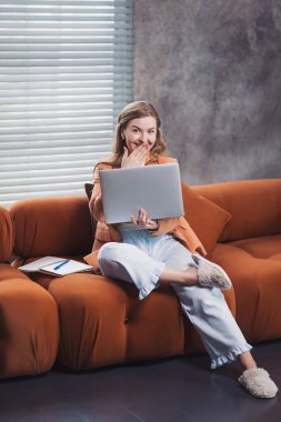 Evde dizüstü bilgisayarla çalışırken rahat bir kanepenin keyfini çıkararak gülümseyen bir serbest yazar. Evden çalışan, iş ceketli, pijama pantolonlu ve terlikli bir kadın.