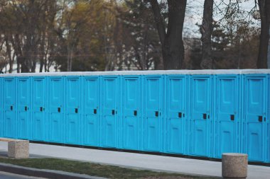 Parlak mavi portatif tuvaletler güneşli bir öğleden sonra bir park ortamında dizilmiş.