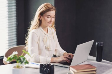 Gündüz saatlerinde modern masadaki dizüstü bilgisayarında kamera ve defterle çalışan genç bir kadın.