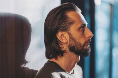 Derin düşüncelere dalmış, bir duvarın yanında duran, yumuşak ışıkta yakalanmış, profilini ön plana çıkaran ve dramatik bir atmosfer yaratan bir birey.