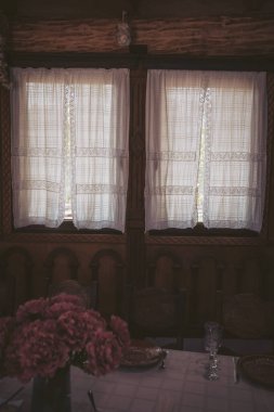 Soft sunlight streams through lace curtains, illuminating a charming dining area with blooming flowers and rustic wooden accents in a cozy home setting