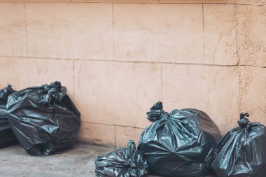 Gündüz saatlerinde kentsel bir alanda siyah çöp torbaları dokulu bir duvara yığılır ve atık yönetimine duyulan ihtiyacı vurgular.