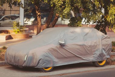 Covered car resting on a city street under the afternoon sun with trees providing shade