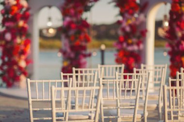 Gün batımında deniz kenarında beyaz sandalyeler ve canlı çiçek süslemeleriyle sersemletici bir açık hava düğün hazırlığı. Parlak renkli çiçekler zarif dizilmiş sandalyeler için çarpıcı bir zemin oluşturur. 