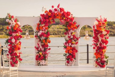 Vibrant floral archway decorates a serene lakeside wedding venue at sunset with colorful blossoms and elegant lanterns