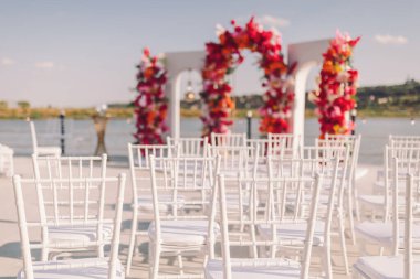 Güneşli bir öğleden sonra, zarif beyaz sandalyeler ve canlı bir çiçek kemeriyle nehir kenarında yapılan açık hava düğün töreni.