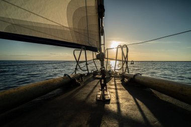 Gün batımında yelkenlinin güvertesinden manzara, teçhizat ve iplerle birlikte. Altın ışık sakin sudan yansıyarak teknenin yapısını belirginleştirir ve bir atmosfer yaratır..