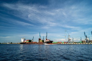 Berrak mavi gökyüzünün altında vinçleri ve konteynırları olan yoğun bir sanayi limanına yanaşmış bir kargo gemisi, sakin suya yansıyor..