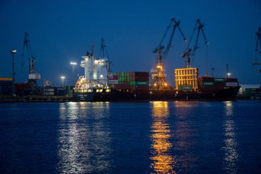 A cargo ship docked at twilight with cranes and stacked containers, reflecting lights on calm water. clipart