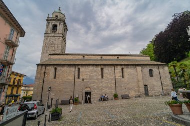 Çan kulesi olan tarihi taştan kilise. Etrafı yeşillik, palmiye ağaçları ve bulutlu bir gökyüzünün altında renkli binalarla çevrili. Ziyaretçiler yakınlarda yürürken ve otururken görülüyor..
