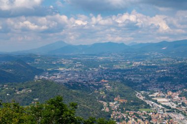 Parçalı bulutlu bir gökyüzünün altında yuvarlanan tepeler ve dağlarla çevrili bir vadi manzarası kentsel alanları, yeşillikleri ve uzak tepeleri gözler önüne seriyor..