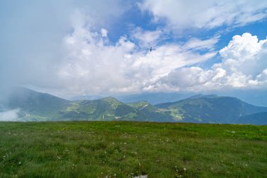Yeşil tepeler, kır çiçekleri ve parçalı bulutlu bir gökyüzünün altında süzülen bir kuşun bulunduğu manzaralı dağ manzarası huzur ve doğal bir ortam sunuyor..