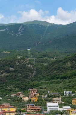 Colorful hillside village surrounded by lush greenery with a cable car station and cables ascending towards the mountain peaks under a partly cloudy sky. clipart