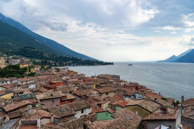 Charming lakeside village with terracotta rooftops, surrounded by green mountains and calm waters under a partly cloudy sky. clipart