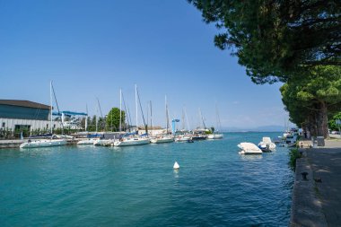 Bereketli yeşillik ve berrak mavi gökyüzü ile çevrili turkuaz suları boyunca rıhtıma yanaşmış yelkenli ve yatları olan manzara marinası. Ağaçlarla kaplı huzurlu bir gezinti alanı..