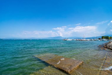 Sakin bir gölün kristal berrak suları sığ kıyıya uzanan küçük bir taş iskelesi var. Yelkenliler ve yatlar, açık mavi gökyüzünün altındaki uzak bir marinaya demirlediler..