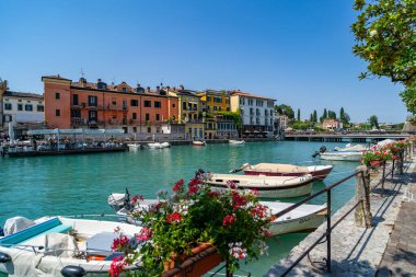 Rıhtım boyunca demirlemiş küçük tekneleri, ön planda canlı çiçek düzenlemeleri ve parlak mavi gökyüzünün altında suyla kaplanmış renkli İtalyan binaları olan büyüleyici kanal manzarası..