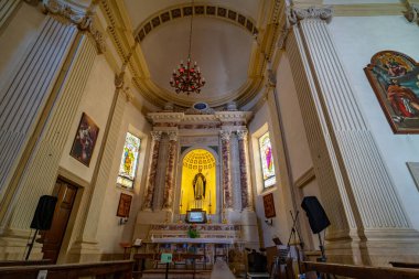 Tarihi bir kilisenin iç manzarası. Karmaşık mimari detaylar, büyük sütunlar ve süslü avizeler sergileniyor. Sunakta dini heykeller, mumlar yer alıyor.