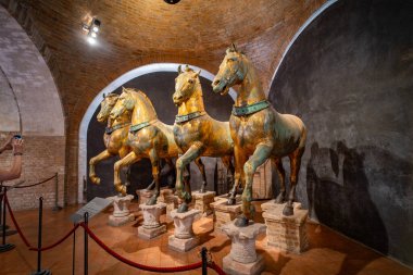 Bir müze sergisinde sergilenen antik bronz at heykelleri, karmaşık işçilik ve tuğla kemerli bir odada sıcak bir aydınlatma altında tarihsel önemi sergiliyor..