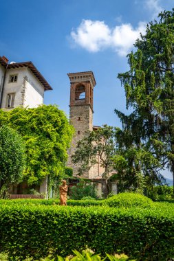 Etrafı yemyeşil bahçelerle çevrili tarihi bir çan kulesi, canlı yeşillikler ve parlak mavi gökyüzünün altında dingin bir heykel..
