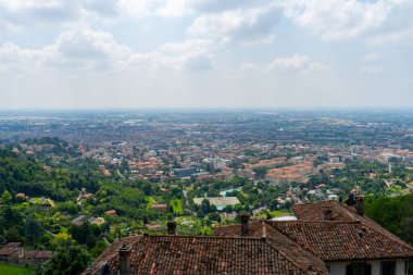 Kentsel gelişimin ve yemyeşil yeşilliğin karışımını yakalayan geniş bir manzara, ön planda terracotta çatıları parçalı bulutlu bir gökyüzünün altında.