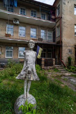 A weathered concrete statue of a figure with missing limbs, standing in an overgrown courtyard in front of an old building with balconies. Ukrain, War, clipart