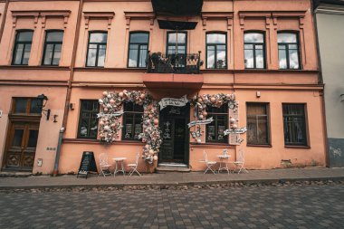 Pastel renkli balonlar ve çiçek düzenlemeleri ile dekore edilmiş rahat bir kafe ön cephesi, parke taşı bir sokakta oturarak tamamlanmış..