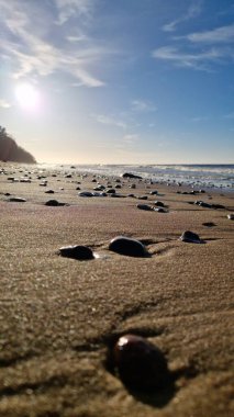 Sahilde parlayan çakıl taşları güneş ışığını yakalar ve arka planda yavaşça dalgalar kırılır..