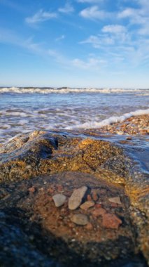 Sahilde büyük, desenli bir kaya. Arkasında hafif dalgalar ve açık mavi bir gökyüzü var..