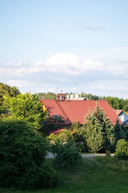 Temiz bir gökyüzünün altında canlı yeşil ağaçların arasına yerleşmiş kırmızı çatılı güzel bir ev manzarası banliyödeki sükunetin özünü yakalıyor..