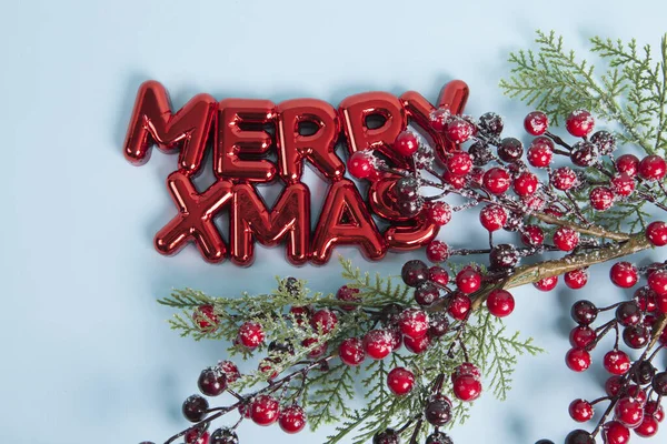メリーXmasというテキストをフィーチャーした光沢のある赤いプラスチック製のクリスマス装飾 写真の上部に赤い果実を塗ったホリーの枝 ブルーの背景 ミニマリスト トレンディな静かな生活写真 — ストック写真