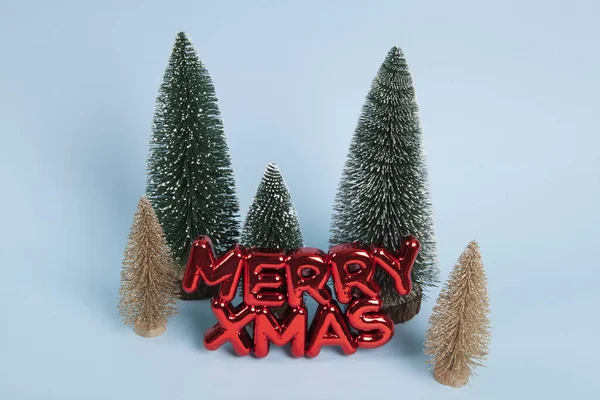 雪の緑と黄金のクリスマスツリーの森の前でメリーXmasを言ったテキストをフィーチャーした光沢のある赤いプラスチックのクリスマスの装飾 ミニマリスト トレンディな静かな生活写真 — ストック写真