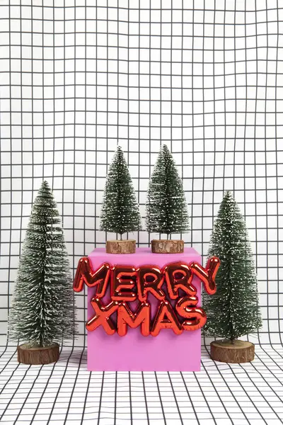 圣诞装饰品一种光滑的红色塑料圣诞装饰品 上面有一段文字写着圣诞快乐 还有一个由几棵小圣诞树和一个黄色立方体组成的小森林场景 其背景是黑色和白色的图形网格 最低限度的静物摄影 — 图库照片