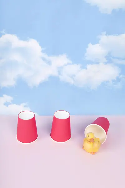 ボノトーのゲームをするために3つの紙コップが使用されます 上向きのカップの下には カップ自体よりもはるかに大きい黄色のアヒルがあります 背景は青空で ピンク色の明るい地面です ロイヤリティフリーのストック画像