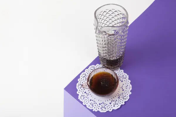 a violet cube made with colored paper to create a 3D optical illusion. On top is a vintage pink coffee cup with a white lace doilyand a glass jug of coffee imitation, fake crystal. Vivid colors and minimal pop art photography.