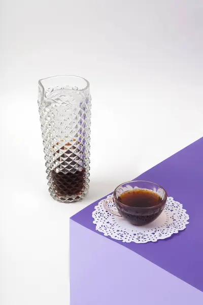 a violet cube made with colored paper to create a 3D optical illusion. On top is a vintage pink coffee cup with a white lace doilyand a glass jug of coffee imitation, fake crystal. Vivid colors and minimal pop art photography.