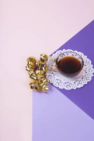 a violet cube made with colored paper to create a 3D optical illusion. On top is a gold Gift ribbon curled near a vintage pink coffee cup with a white lace doily. Vivid colors and minimal pop art photography.